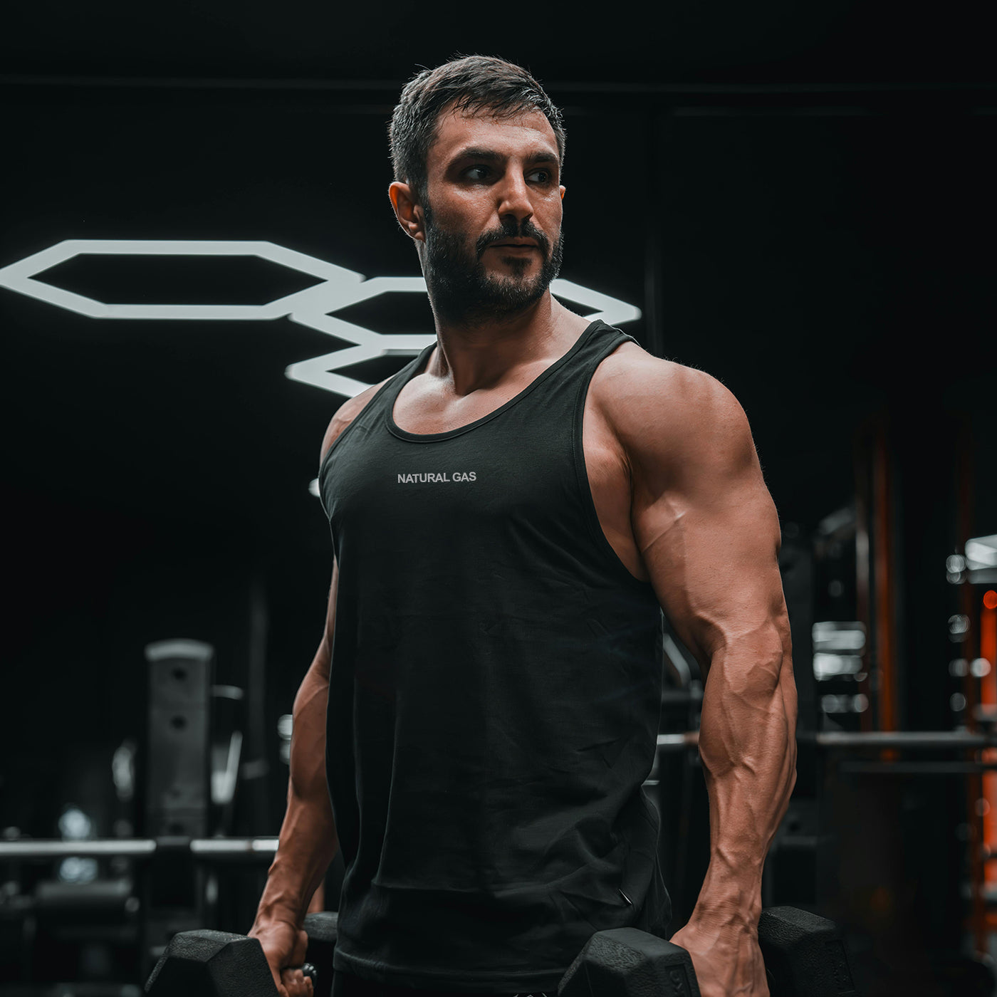 Muscular male athlete wearing Natural Gas black tank top lifting dumbbells in a gym – fitness apparel for performance and style
