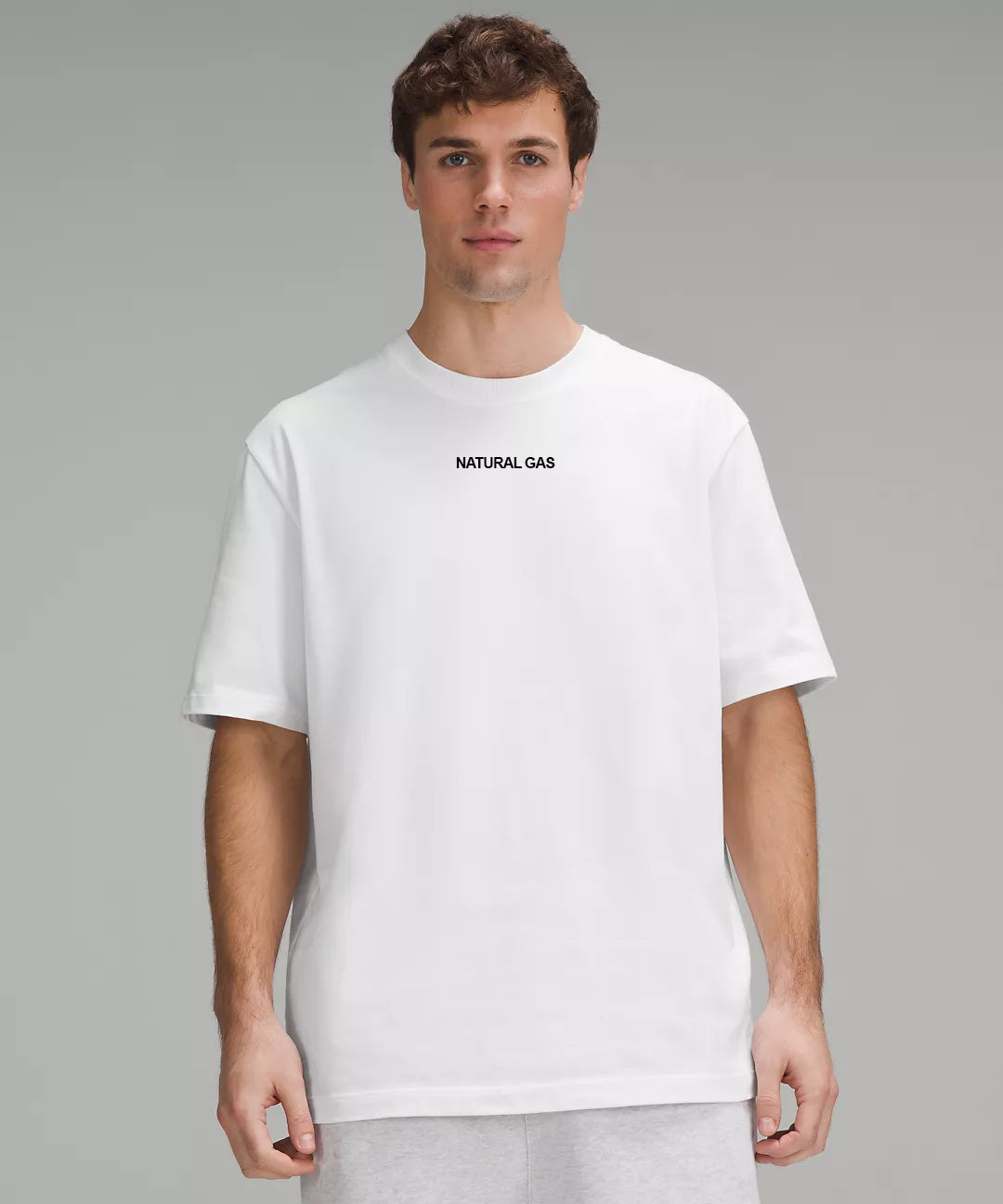 A man wearing a white crew neck t-shirt with a black printed 'Natural Gas' logo on the chest and a woven side tag detail.