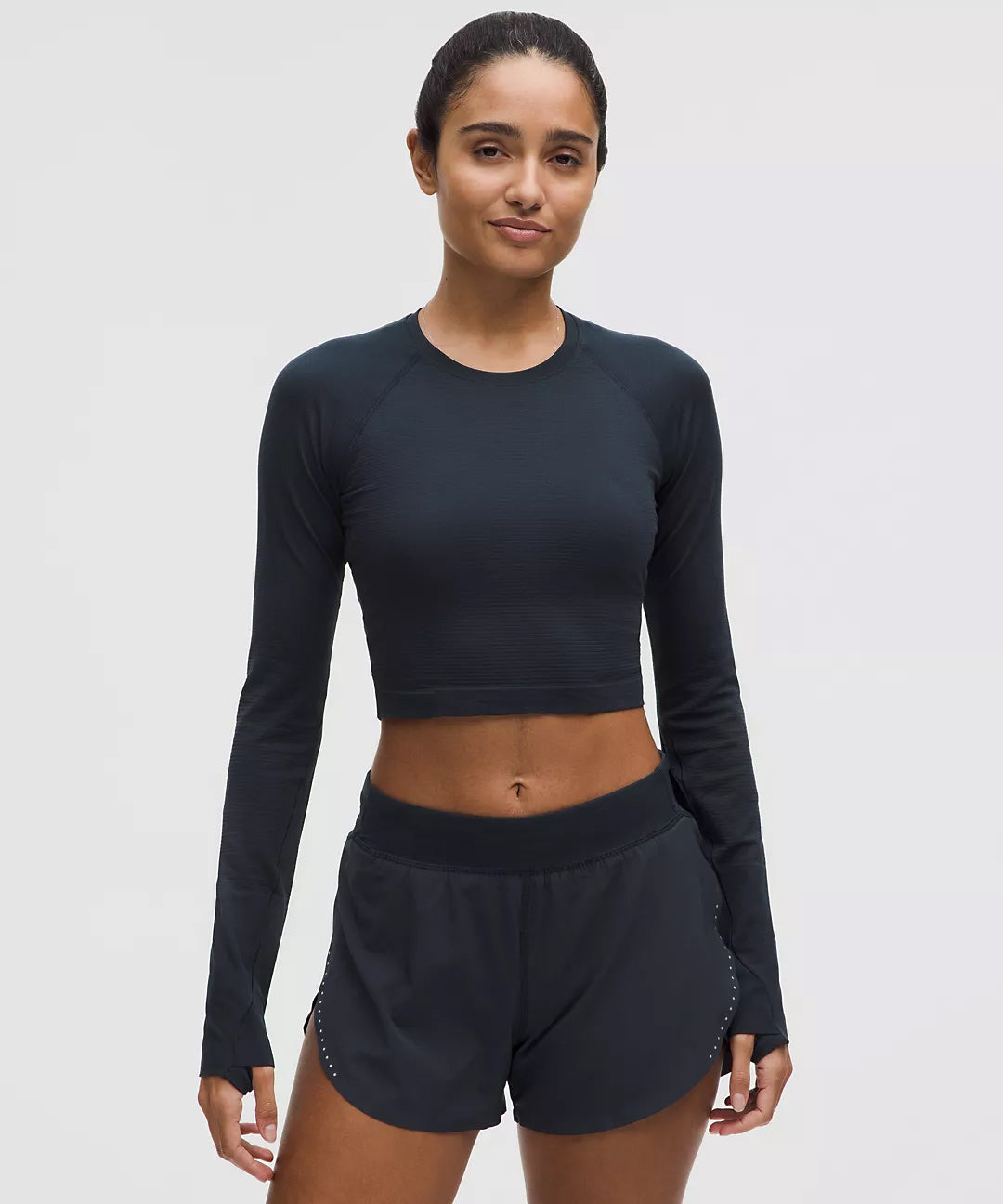 Woman wearing a fitted black long sleeve crop top and black athletic shorts, front-facing studio shot showcasing sleek gym apparel designed for performance and comfort.